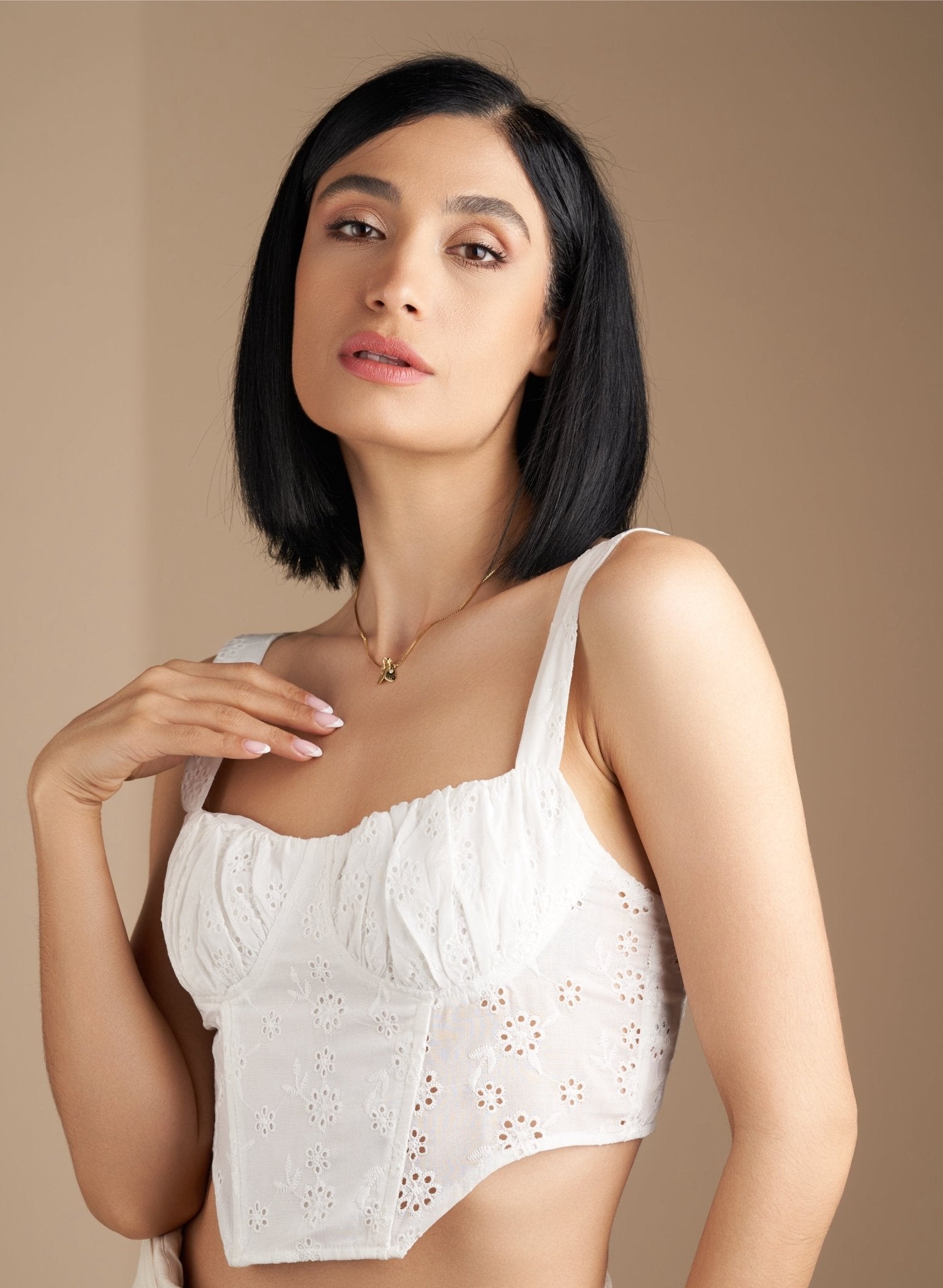 Model wearing a white embroidered top and a delicate gold necklace, posing against a neutral background